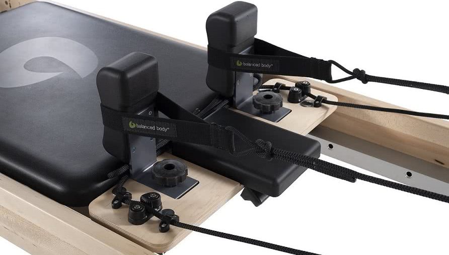 Pilates reformerio platforma su reguliuojamais peties atramomis ir juodu apmušalu.